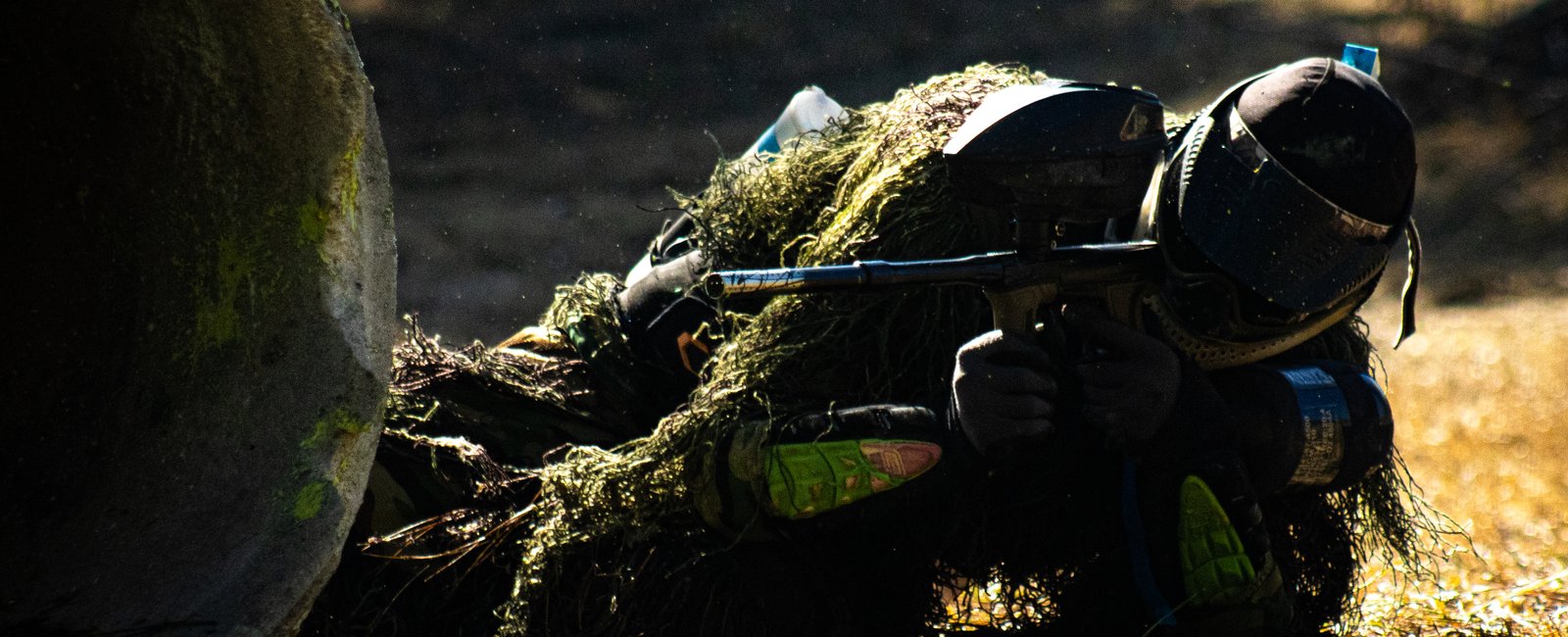 Ghillie-suited player prone behind a concrete pipe bunker in the woods